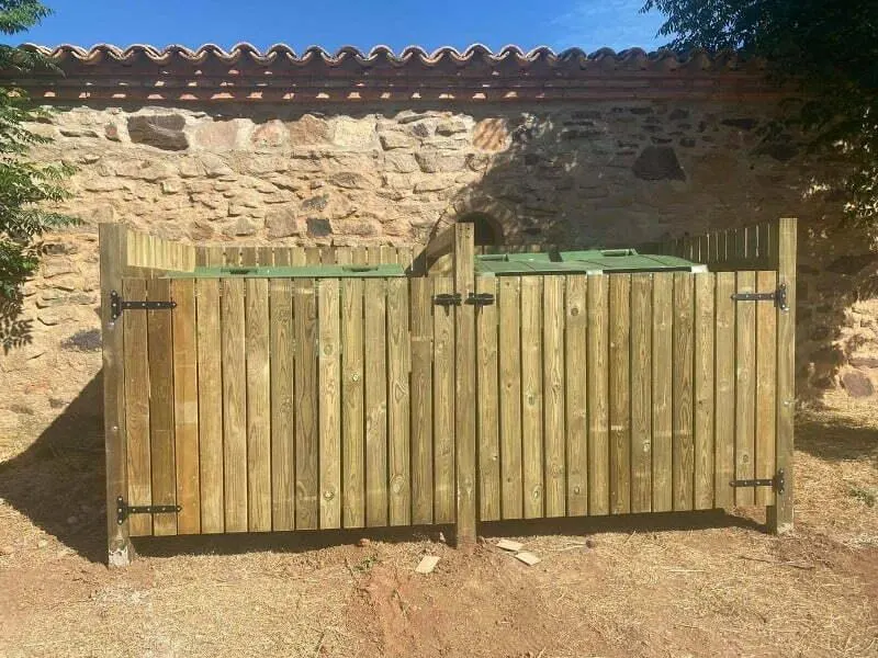 Cubrecontenedor de madera sin tejado para dos contenedores junto a un muro de piedra