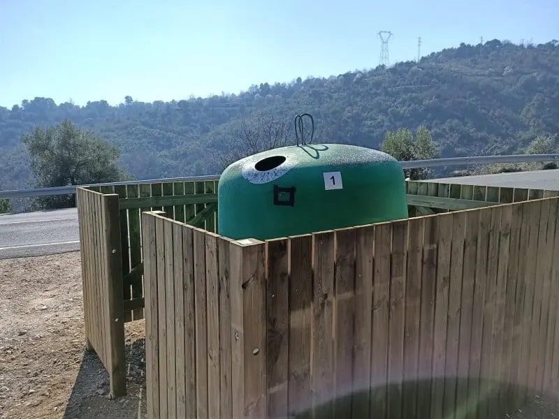 Cubrecontenedor de madera sin tejado que rodea un iglú de vidrio verde, con montes al fondo