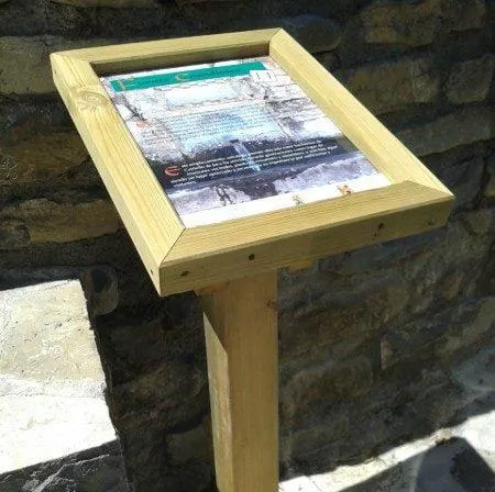 Atril interpretativo de madera con panel informativo junto a un muro de piedra